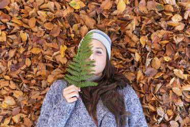 Beautiful woman lying down on autumn leaves while covering face with fern leaf in Cannock Chase woodland - WPEF03678