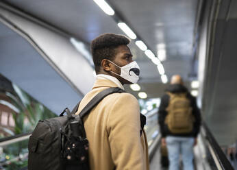 Junger Mann mit Gesichtsmaske steht auf einer Rolltreppe im Bahnhof - JCCMF00015