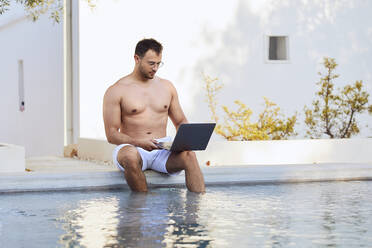 Young man working on laptop while sitting with legs dangling in swimming pool - UKOF00080