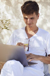 Junger Mann hält ein Glas Wasser in der Hand und arbeitet an einem Laptop an der Wand - UKOF00092