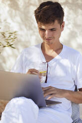 Young man holding glass of water while working on laptop against wall - UKOF00092