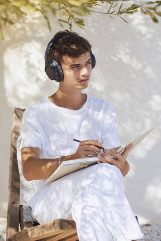 Thoughtful young man with book and pen listening music through headphones against white wall stock photo