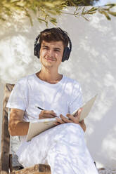 Lächelnder Mann, der beim Hören von Musik an einer weißen Wand im Hinterhof nachdenkt - UKOF00094