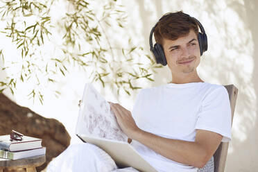 Lächelnder junger Mann mit Buch, der wegschaut, während er über Kopfhörer im Hinterhof Musik hört - UKOF00096