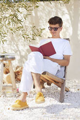 Junger Mann sitzt auf einem Stuhl und liest ein Buch an der Wand im Hinterhof - UKOF00101