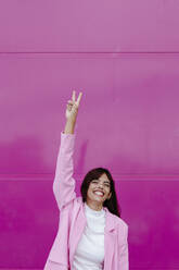 Smiling woman with hand raised doing peace sign while standing against pink wall - TCEF01332