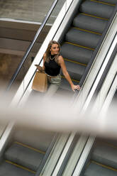 Frau mit Einkaufstasche und Mobiltelefon schaut weg, während sie auf einer Rolltreppe steht - MAUF03623