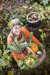 Lächelnde Frau, die einen Obst- und Gemüsekorb trägt, während sie im Garten steht - HHF05599