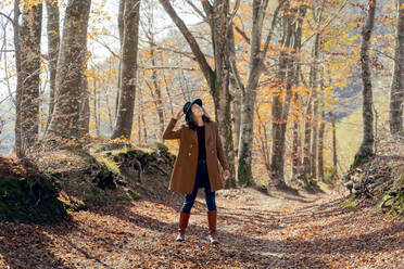 Frau mit Hut und Jacke, die auf einem Fußweg im Wald steht und nach oben schaut - FMOF01282
