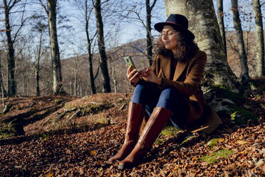 Frau benutzt Mobiltelefon, während sie unter einem Baum im Wald sitzt - FMOF01286