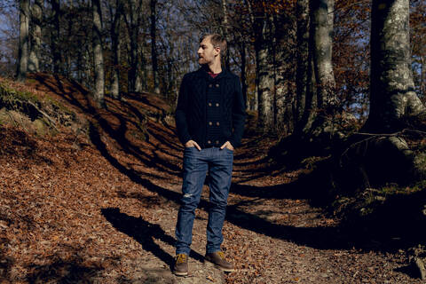 Man looking away while standing with hands in pockets on forest path stock photo