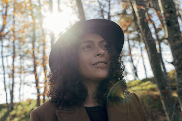 Curly hair woman wearing hat looking away while exploring forest - FMOF01308