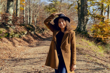 Smiling woman wearing hat and jacket standing on forest path - FMOF01315