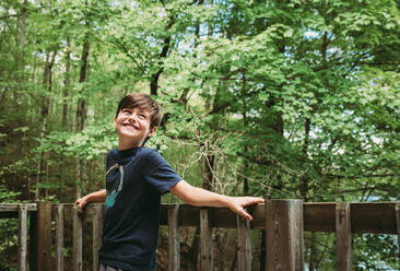 Happy young boy leaning against deck railing with trees in background. - CAVF90651