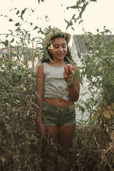 A smiling girl looks at a tomato in her small garden at home - CAVF90747