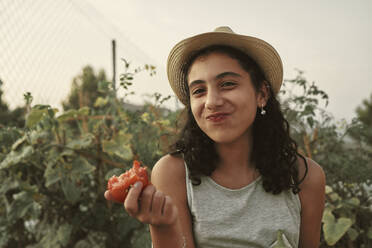 Ein Mädchen mit lockigem Haar isst eine Tomate in ihrem eigenen Garten - CAVF90753