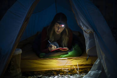 Young woman writing in diary in tent at night - CAVF90787