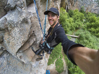Smiling man taking selfie while climbing rocky cliff - CAVF90792