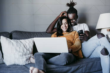 Happy young man with pregnant woman looking at mobile phone while using laptop at home - EBBF01595