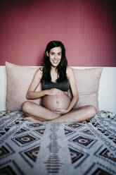 Lächelnde schwangere Frau sitzt auf dem Bett gegen die rote Wand zu Hause - EBBF01604