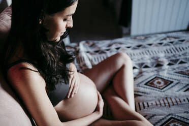 Schwangere Frau berührt den Bauch, während sie zu Hause auf dem Bett sitzt - EBBF01605