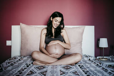 Glückliche schwangere Frau, die ihren Bauch betrachtet, während sie im Schlafzimmer zu Hause sitzt - EBBF01606