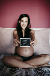 Mid adult pregnant woman showing ultrasound scan while sitting on bed at home - EBBF01607