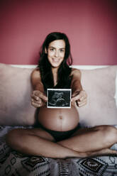 Schwangere Frau im mittleren Erwachsenenalter zeigt Ultraschalluntersuchung, während sie zu Hause auf dem Bett sitzt - EBBF01607