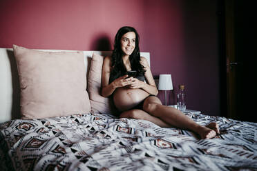 Lächelnde schwangere Frau, die wegschaut, während sie im Schlafzimmer zu Hause ein Mobiltelefon benutzt - EBBF01608