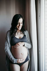 Smiling pregnant woman with hands in stomach standing by window at home - EBBF01609