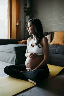Nachdenkliche schwangere Frau hält den Bauch, während sie zu Hause auf einer Übungsmatte sitzt - EBBF01614