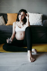 Lächelnde schwangere Frau mit einem Glas Trinkwasser auf einer Matte im Wohnzimmer zu Hause sitzend - EBBF01620