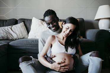 Junger Mann mit schwangerer Frau, die zu Hause ihren Unterleib betrachtet - EBBF01629