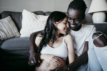 Lächelnder junger Mann mit schwangerer Frau, die zu Hause im Wohnzimmer sitzt - EBBF01631