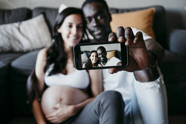Lächelnder junger Mann mit schwangerer Frau macht Selfie zu Hause - EBBF01633