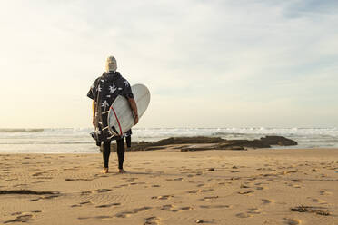 Mann hält Surfbrett auf Sand und schaut auf das Meer gegen den Himmel - KBF00622