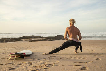 Männlicher Surfer ohne Hemd, der sich auf dem Surfbrett am Strand ausstreckt - KBF00626
