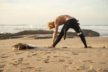 Shirtless male surfer exercising by surfboard at beach - KBF00628