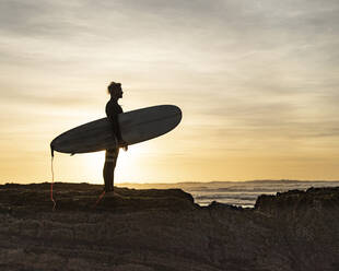 Silhouette Mann trägt Surfbrett auf Felsformation am Strand bei Sonnenuntergang - KBF00633