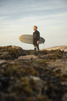 Mann stehend mit Surfbrett auf Felsformation am Strand gegen Himmel - KBF00635