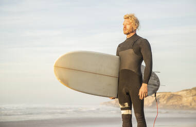 Blonder Mann im Neoprenanzug, der ein Surfbrett trägt, während er am Strand gegen den Himmel schaut - KBF00637