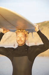 Lächelnder blonder Mann mit Surfbrett über dem Kopf am Strand bei Sonnenuntergang - KBF00641