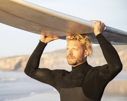 Männlicher Surfer trägt Surfbrett über dem Kopf am Strand bei Sonnenuntergang - KBF00642