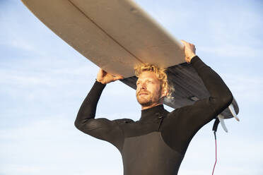 Blonder männlicher Surfer trägt Surfbrett über dem Kopf am Strand gegen den Himmel - KBF00643