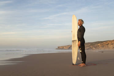 Surfer schaut weg, während er mit seinem Surfbrett am Strand gegen den Himmel steht - KBF00646