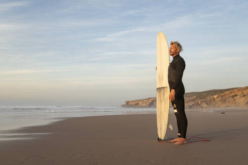 Surfer schaut weg, während er mit seinem Surfbrett am Strand gegen den Himmel steht - KBF00646