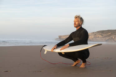 Blonder Surfer mit Surfbrett auf dem Schoß hockt am Strand gegen den Himmel - KBF00647