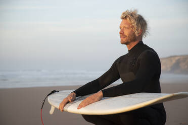 Blonder Mann mit Surfbrett auf dem Schoß hockt am Strand gegen den Himmel - KBF00648