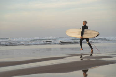 Surfer läuft mit Surfbrett gegen Wellen im Meer bei Sonnenuntergang - KBF00649