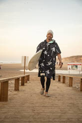 Mittlerer erwachsener Mann mit Surfbrett auf der Strandpromenade gegen den Himmel - KBF00650